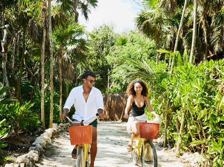 Wide shot of smiling couple riding bikes to beach at tropical resort