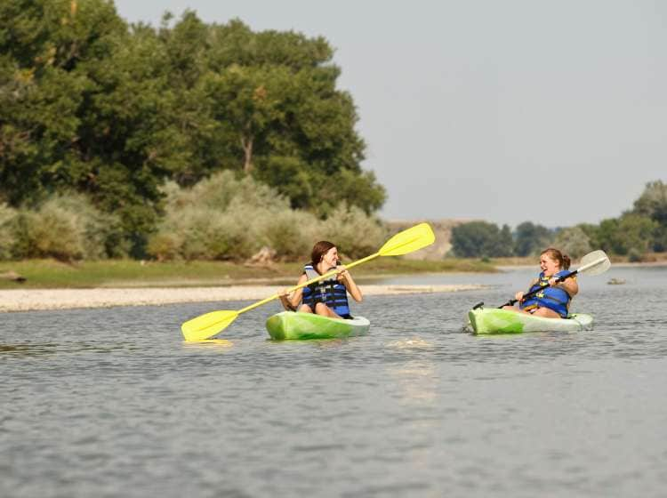 Two girls in life jackets laugh and smile at each other while paddling their green kayaks along the Yellowstone River in Eastern Montana.