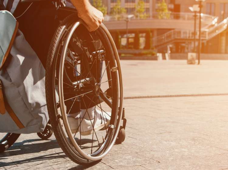 cropped panoramic view of man using wheelchair with bag on street with sunlight