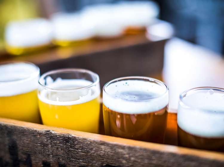 Flight of four beers lined up on a wooden table, with selective focus. Photo taken outdoors with natural lighting; beers range in color from light to very dark. Background out of focus.