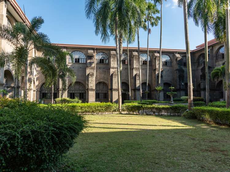 Manila, Philippines - March 5, 2019: San Augustin convent. Green garden as courtyard with brown walls under evening sun, green palm trees and bushes under blue sky.