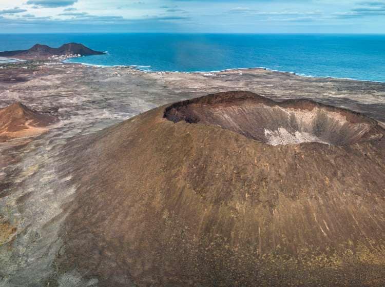 Aerial view of a coastal volcanic landscape (Viana volcano, Sao Vicente) with a distinct crater in the foreground, sparse vegetation, and a small village by the sea, under a cloudy sky