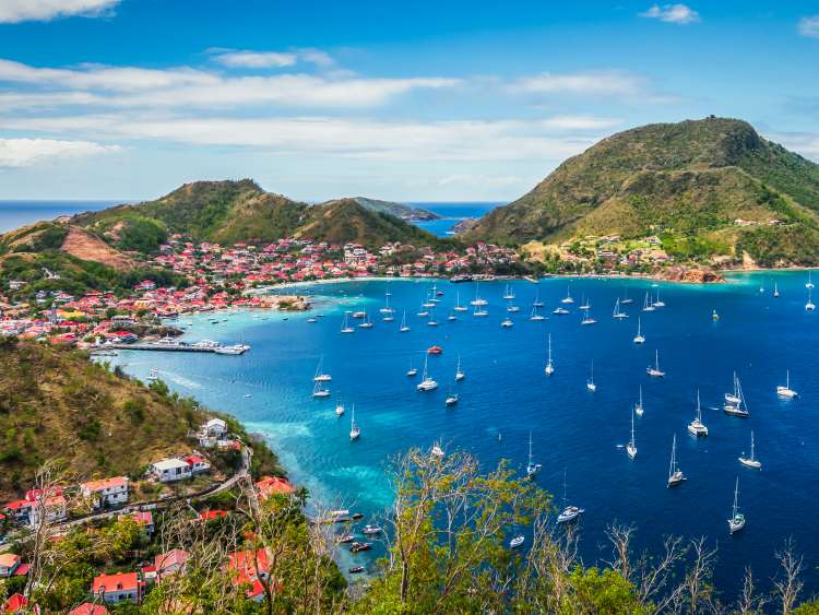 Colorful image of Guadeloupe Terre de Haut bay and town with buildings along the coastline. Small ships anchored in port. Cruise destination. Green mountains in the background. Blue sky and some white clouds.