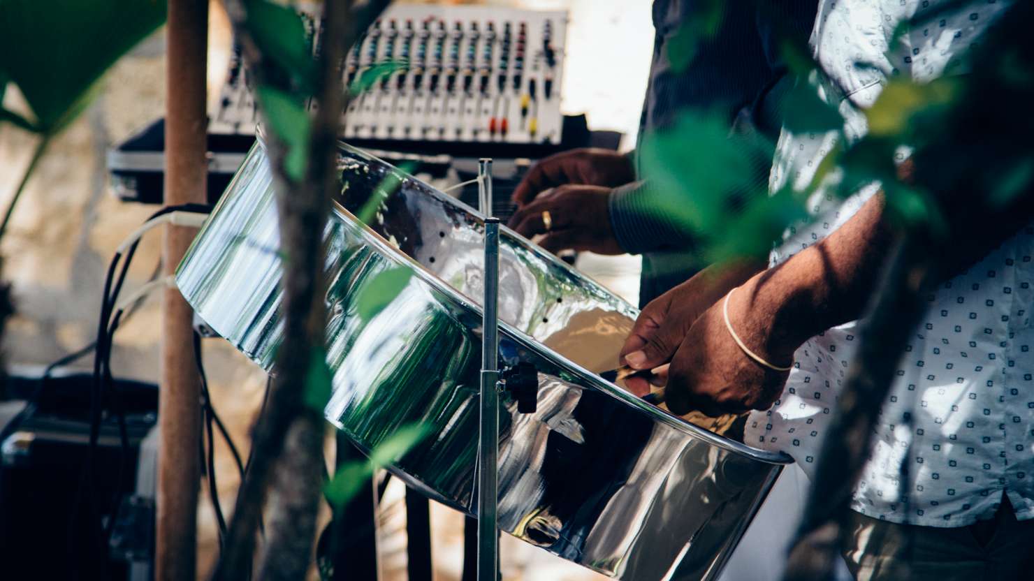 A set of steel drums are played on stage at a live event. Greenery can be seen in the foreground.