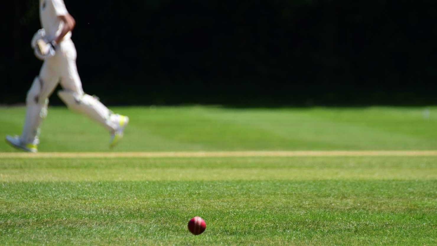 A cricket ball in the foreground on a grass field with a cricket player running on the pitch in the background