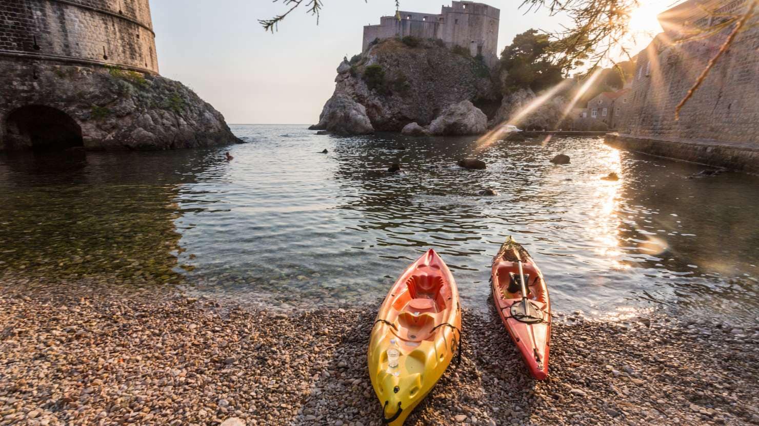 Two kayaks on the shoreline of Kolorina in Dubrovnik, a small rocky inlet between fortresses at sunset.