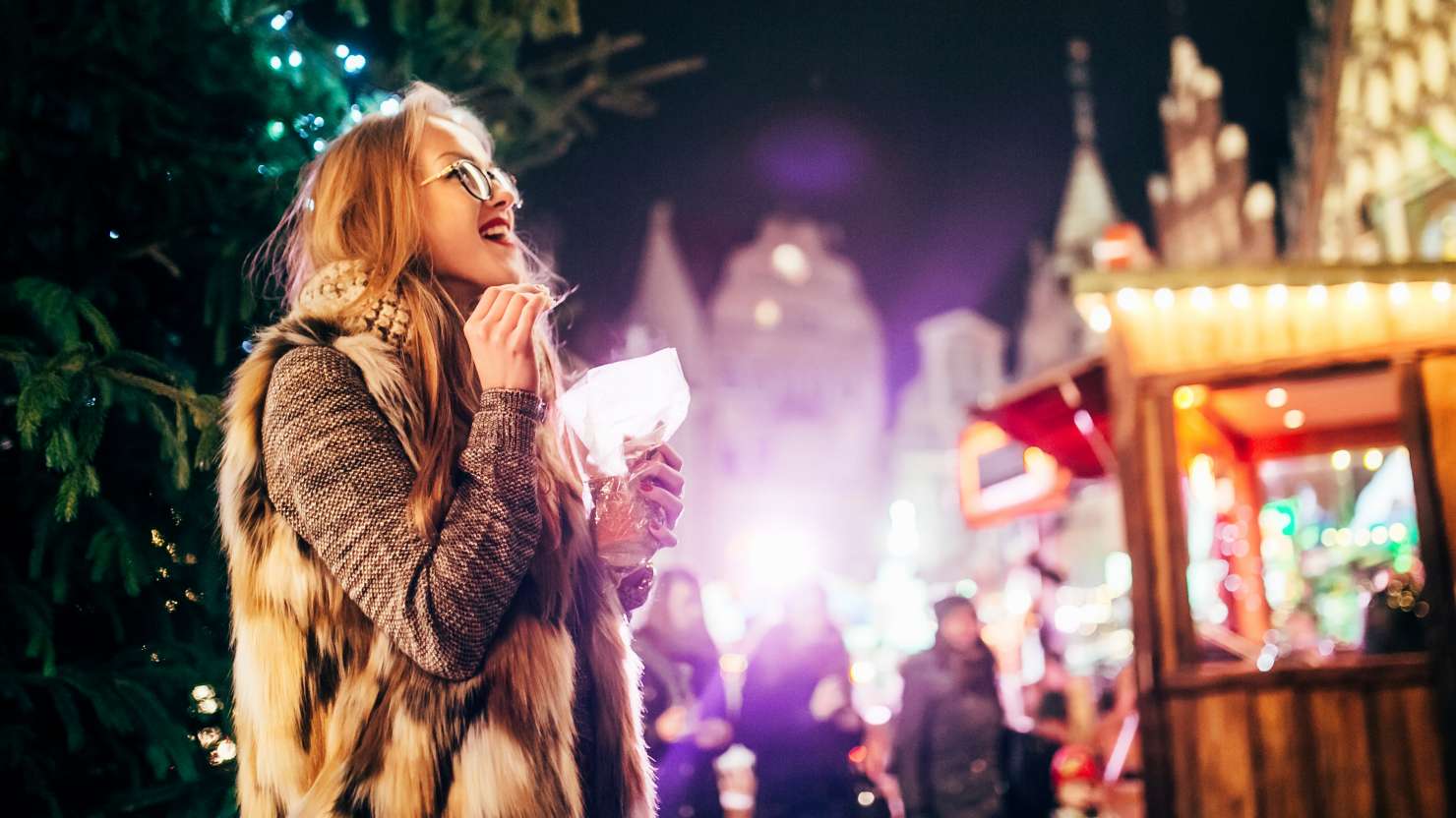 Street portrait of smiling beautiful young woman with glasses on the festive Christmas fair. Model eat market goods at camera and look in sky.  Christmas market