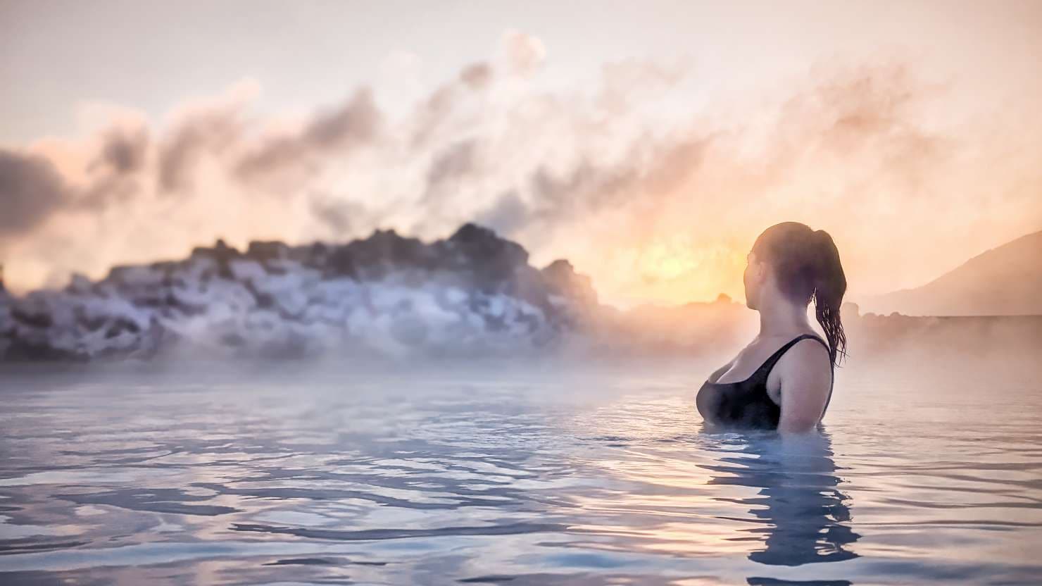 A woman with her hair tied back enjoys the warm waters of a hot spring in Iceland as the sun sets behind misty rocks. The sky glows with romantic colors, creating a serene and tranquil atmosphere around her.