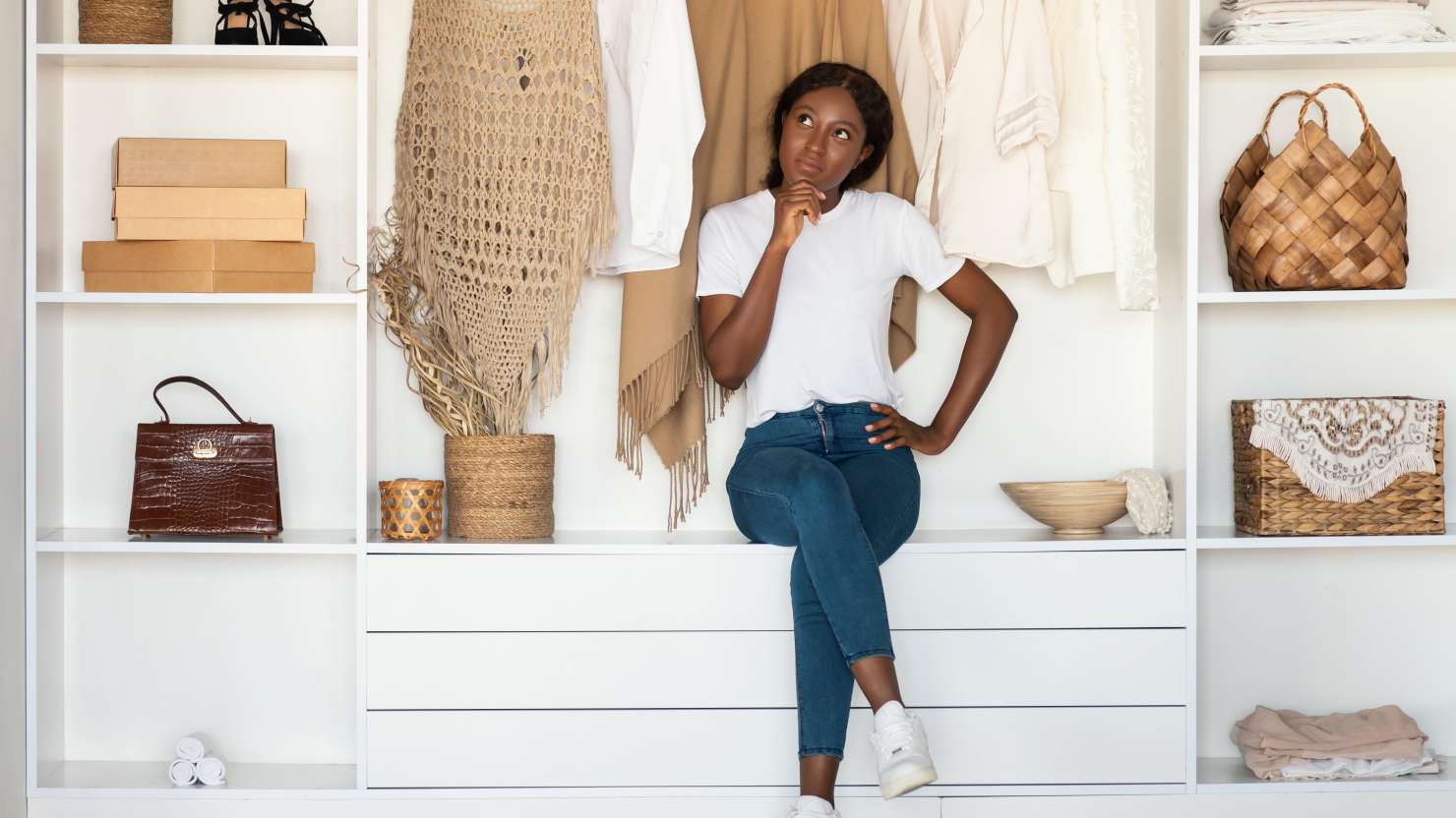 Pensive Black Female Thinking Among Clothes Sitting In Wardrobe At Home. Full Length Shot Of Thoughtful Shopaholic Lady. Fashion Blogging, Style And Outfit Ideas Concept