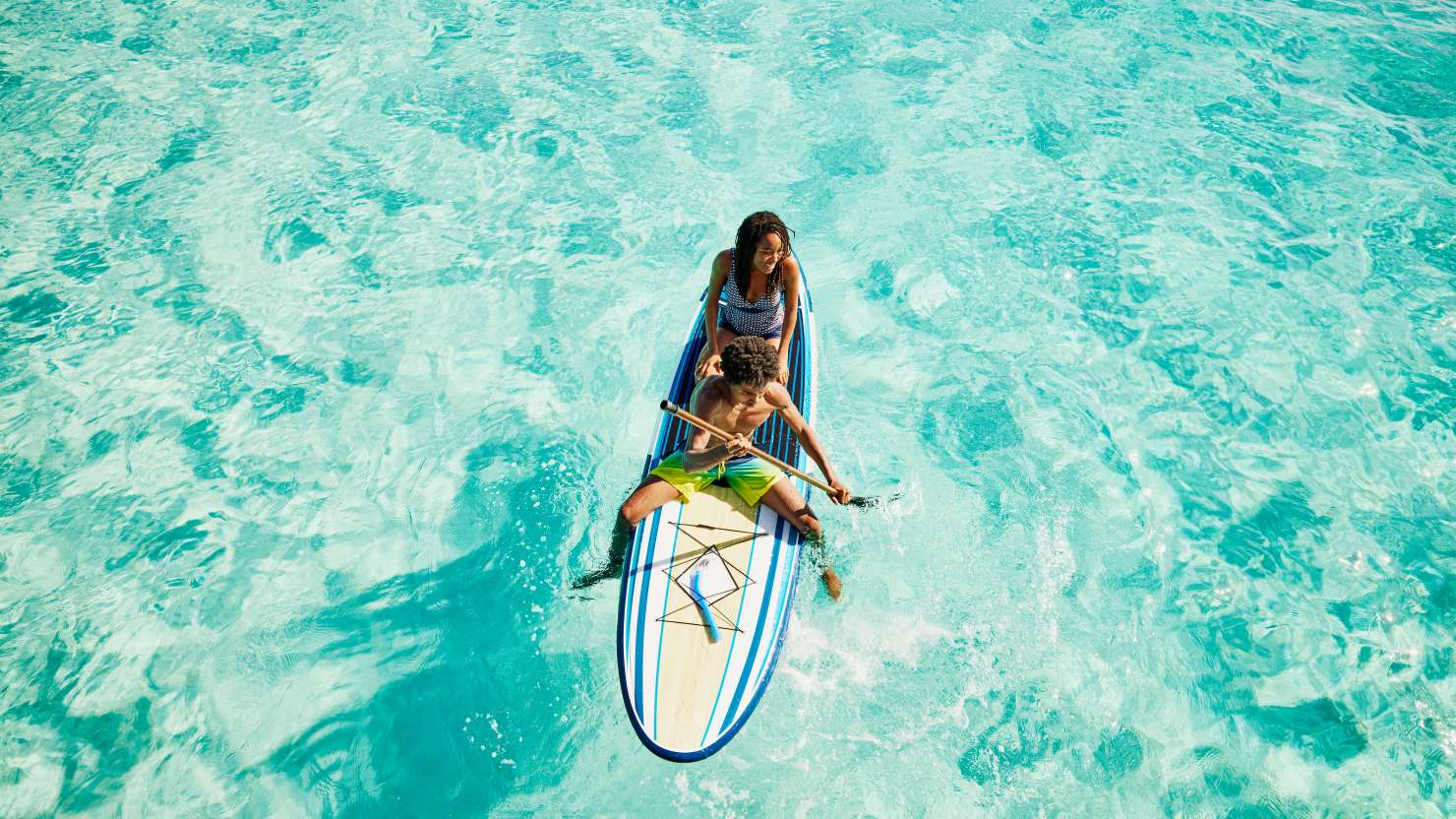 Wide shot of teenage brother and sister paddling paddleboard in ocean during family vacation in Caribbean Sea