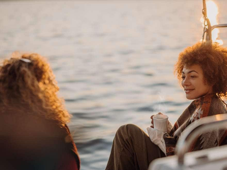 Two women with blond and brown curly hair having a conversation while sitting on boat deck in the morning with blankets and cups of warm drinks, looking at each other with smiling facial expression, rear view